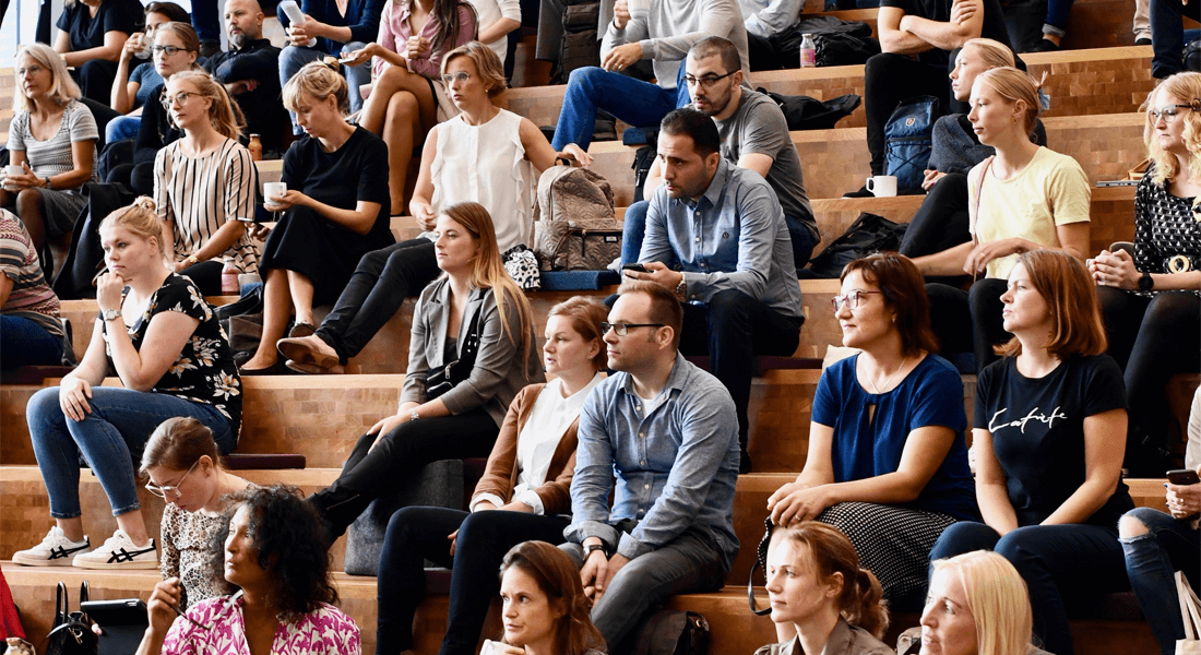 En stor flok voksne mennesker er til efteruddannelse på Københavns Universitet og sidder på trapper i et auditorium og kigger i en retning , imens de lytter til et oplæg