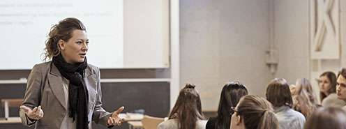 Kvindelig forelæser med krøllet hår i hestehale står op og underviser en flok studerende, der sidder ned og kigger på underviseren